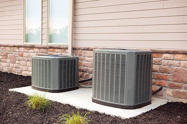 Two air conditioning units on a concrete pad next to a building with beige siding and a stone base.