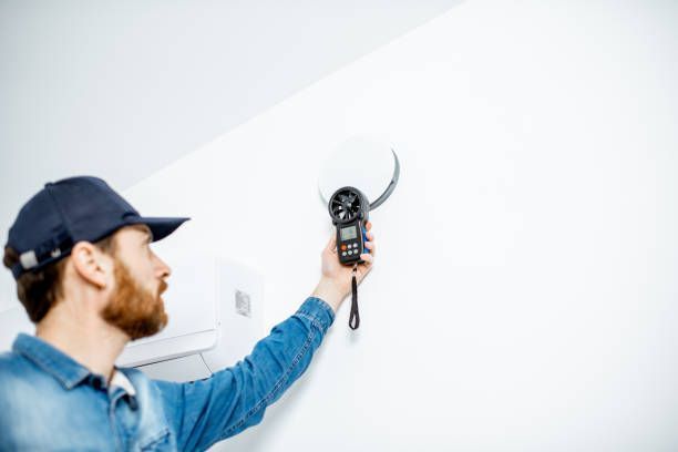 Man in blue shirt and cap uses a digital anemometer on a white vent.