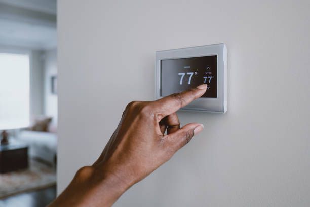 A person's hand adjusting a digital thermostat on a white wall. The temperature is 77 degrees.