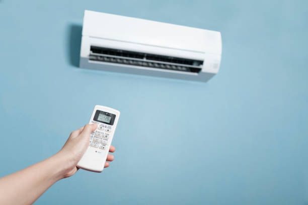 Hand holding a white remote, pointing towards a white air conditioner unit mounted on a light blue wall.