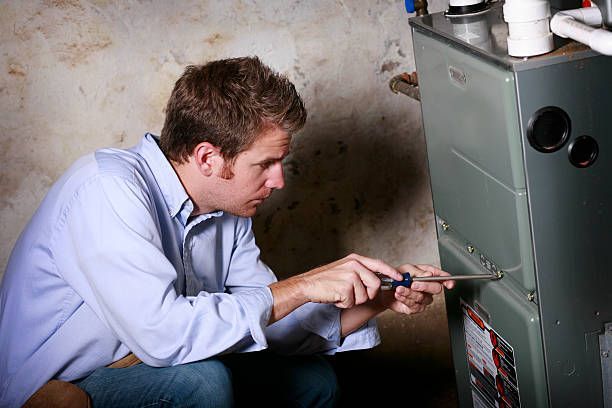 Man in a blue shirt using a screwdriver on a furnace in a basement.