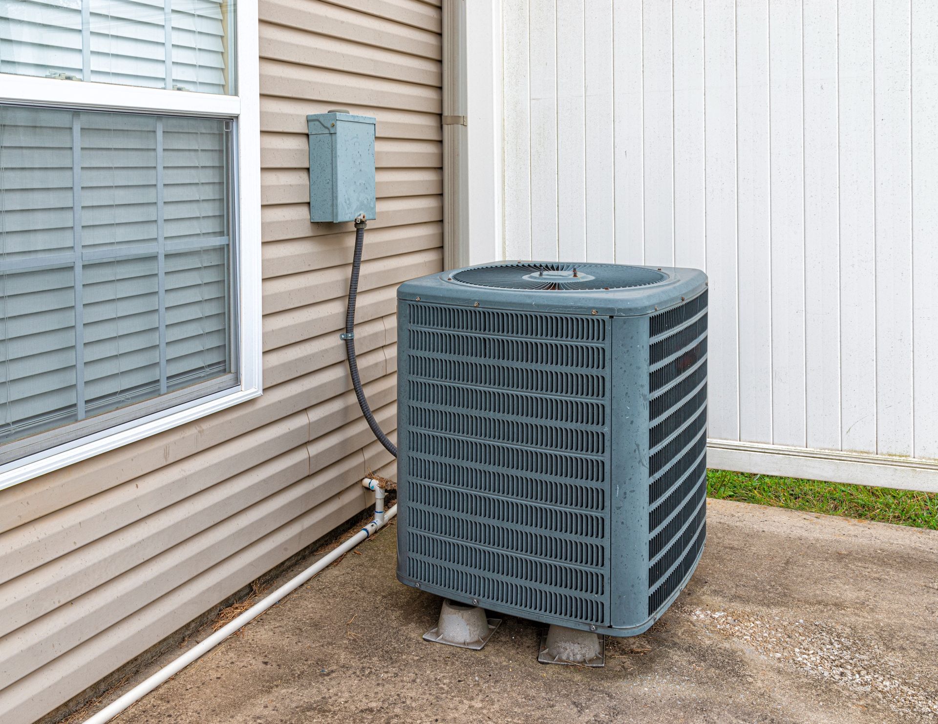 Air conditioning unit outside a building near a window and white fence.