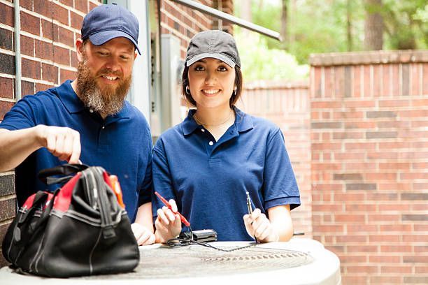 Two HVAC technicians, smiling, prepare to service an AC unit outside a brick building.