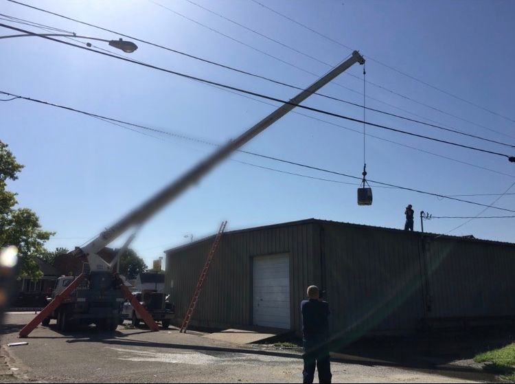 Crane lifting a box over a building near power lines on a sunny day. People on the roof and ground watch.