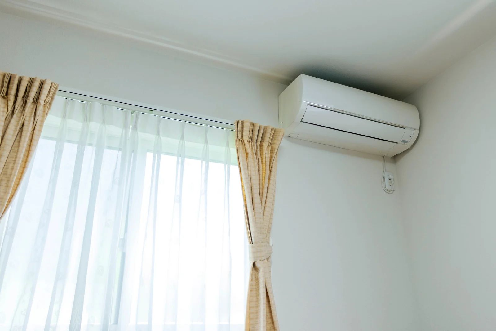 White wall-mounted air conditioning unit above a window with cream curtains.