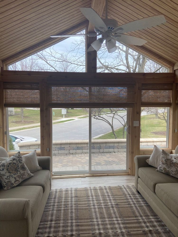 Sunroom with beige couches, plaid rug, wooden accents, large windows, and a ceiling fan.