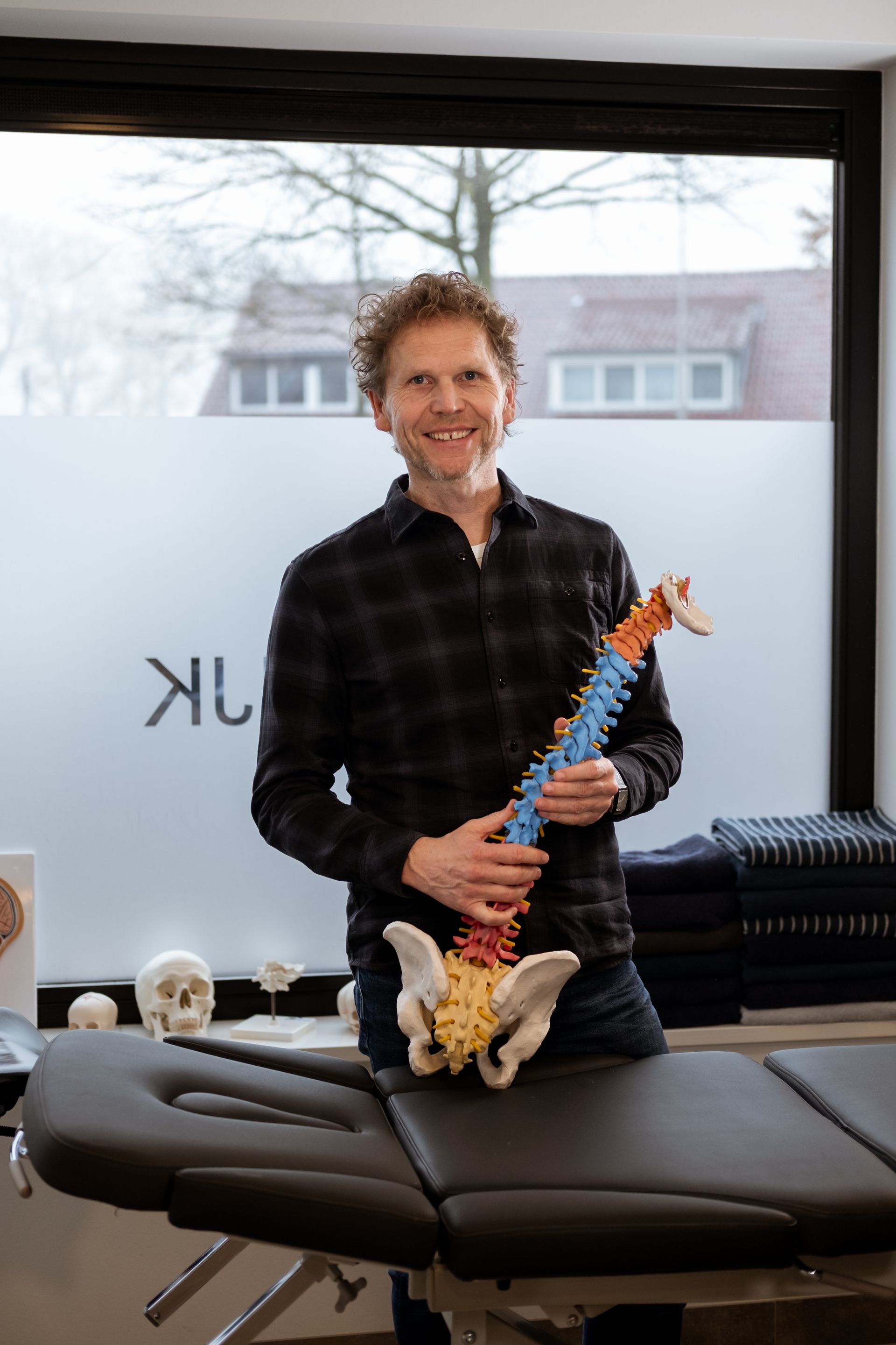 Man in roze shirt en blauwe spijkerbroek aait een zwart-witte hond in een veld met paarse bloemen.