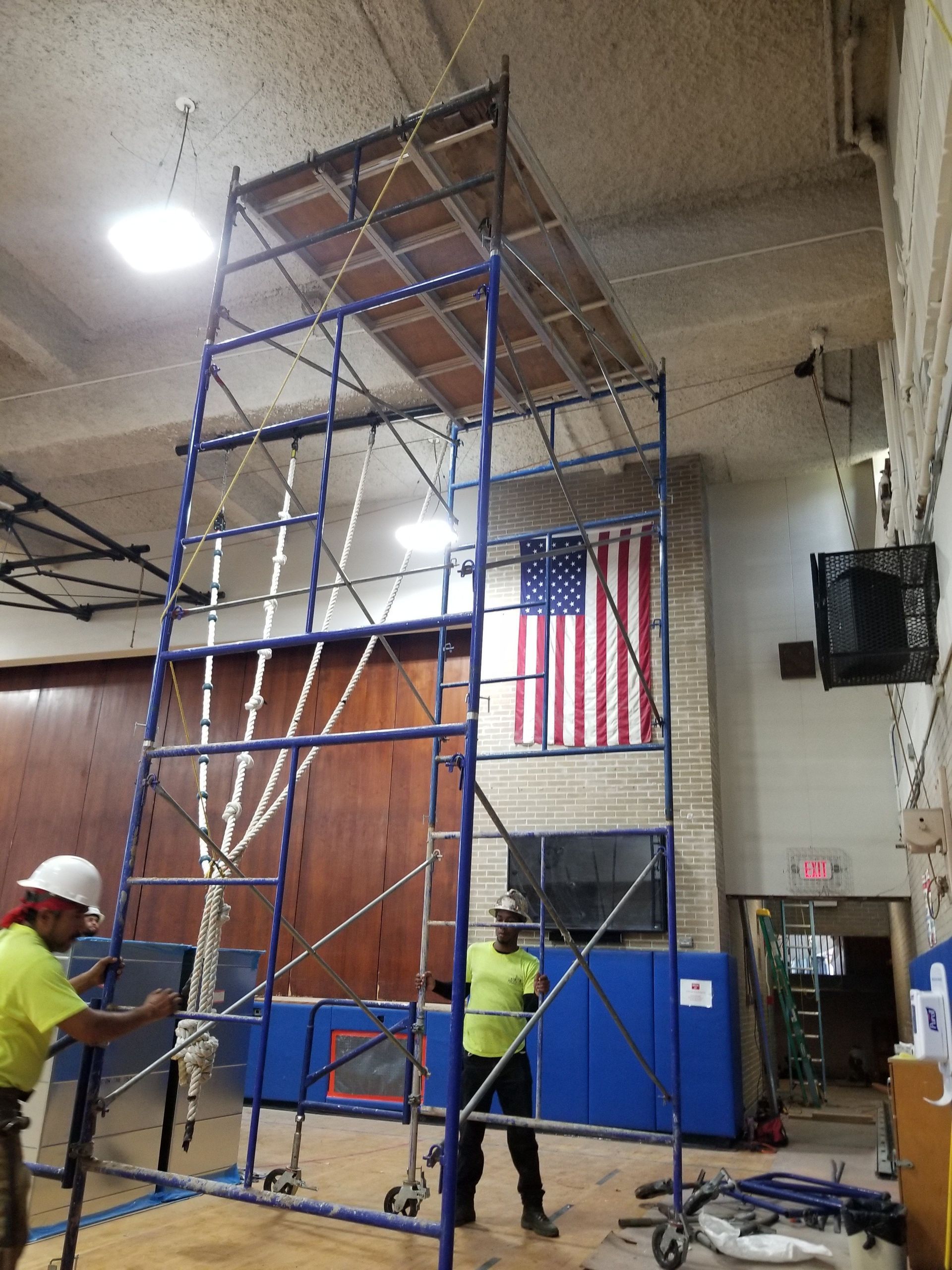 Two workers assembling a tall blue scaffold in a gym, with an American flag in the background.