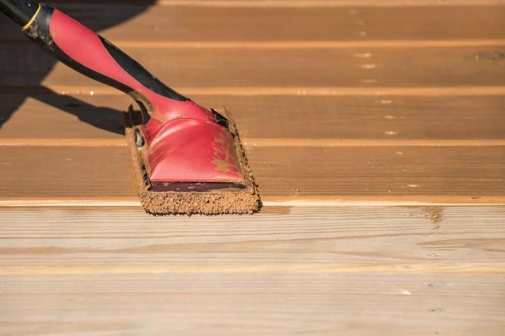 Red and black handled brush applying stain to a wooden deck.