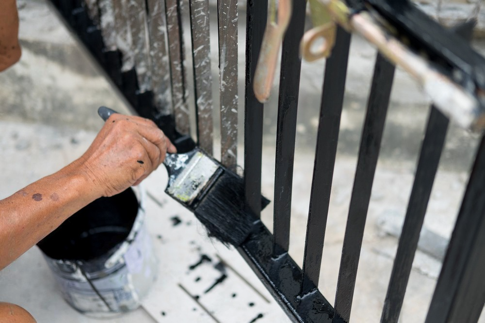 Person painting a black metal fence with a paintbrush, outdoors.