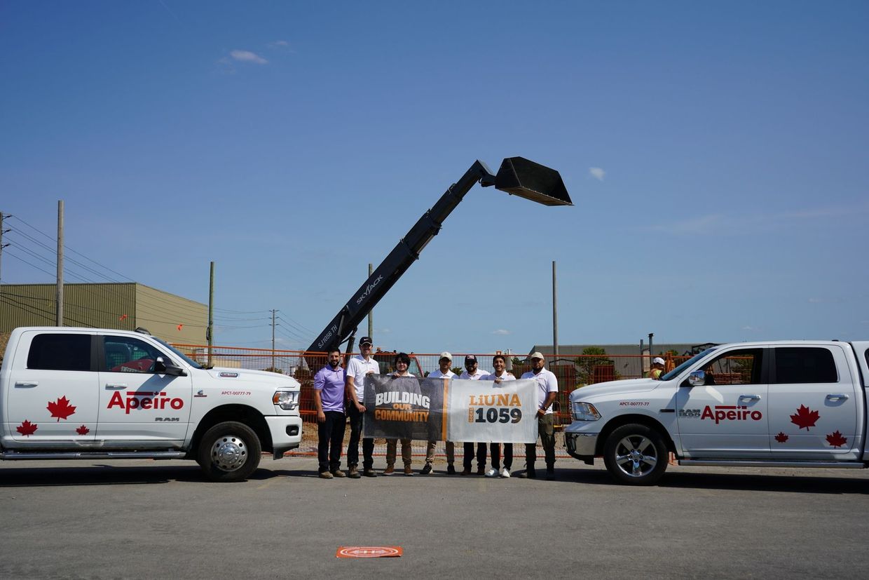 A group stands between two white pickup trucks with Apeiro logos, holding banners in a construction yard with a crane.