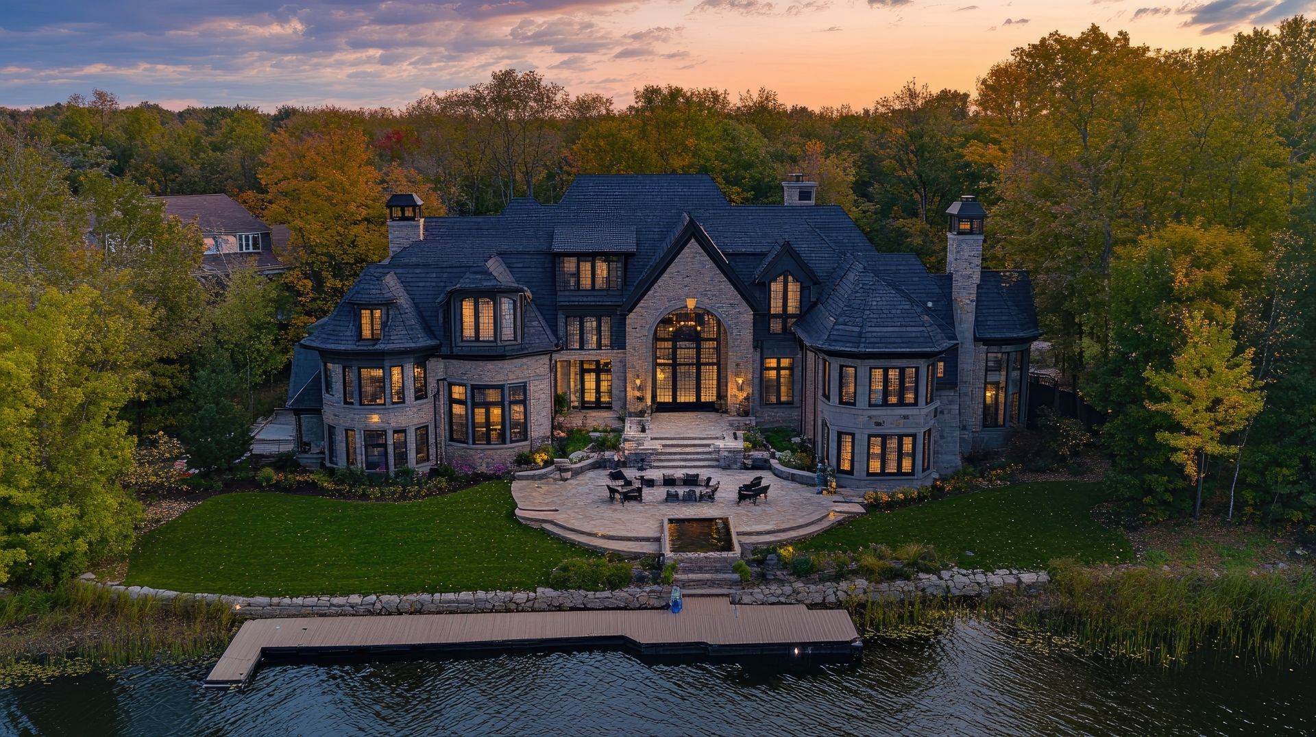 Large stone house on a lake at sunset with a patio and dock surrounded by trees.