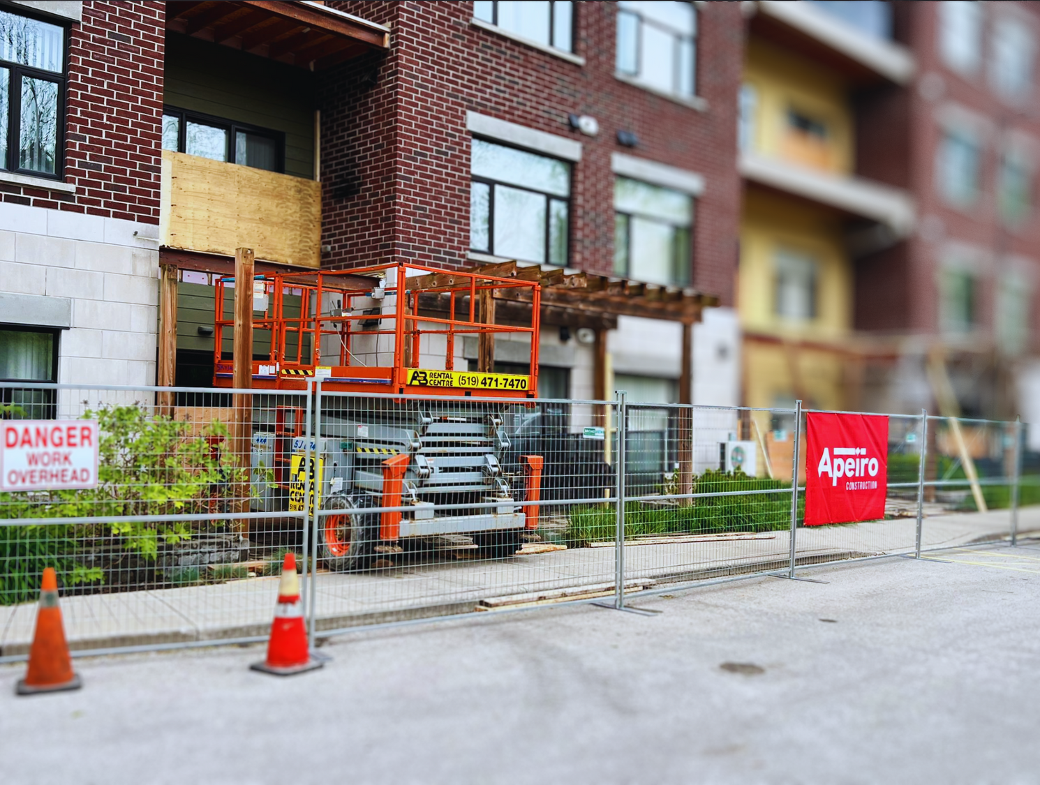 Construction site in front of a brick apartment building, with fencing, cones, and a truck lift.