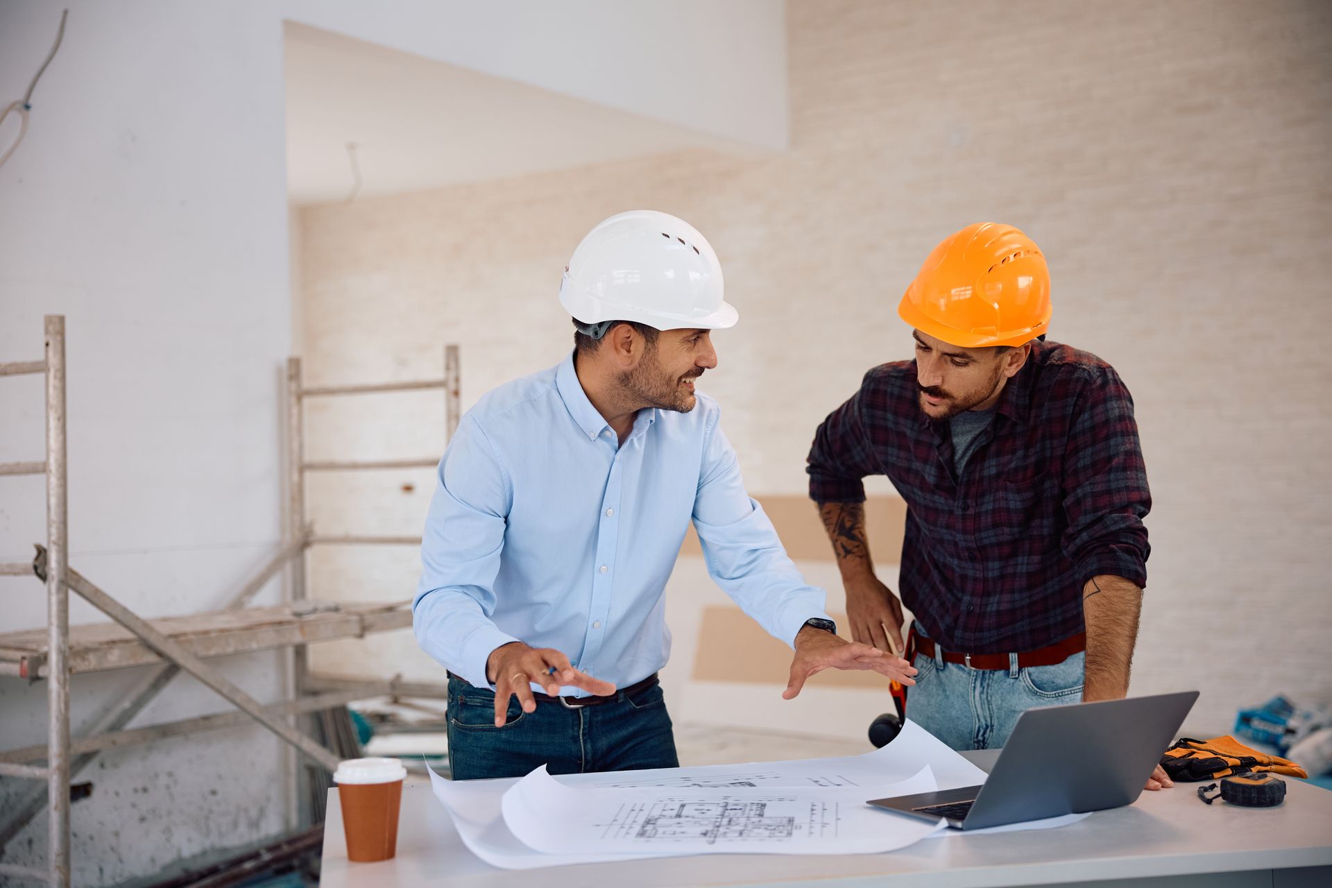 Two construction workers are looking at a blueprint and a laptop.