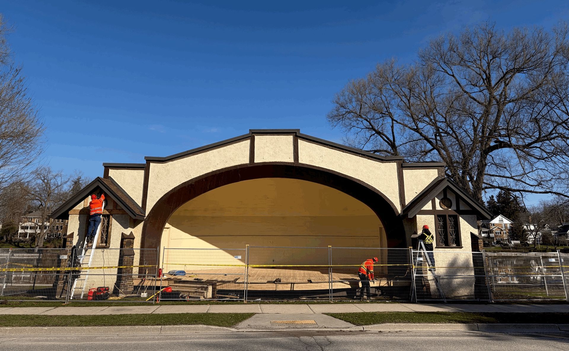 Outdoor band shell stage with tan arch, red accents, and a blue sky above a park fence.