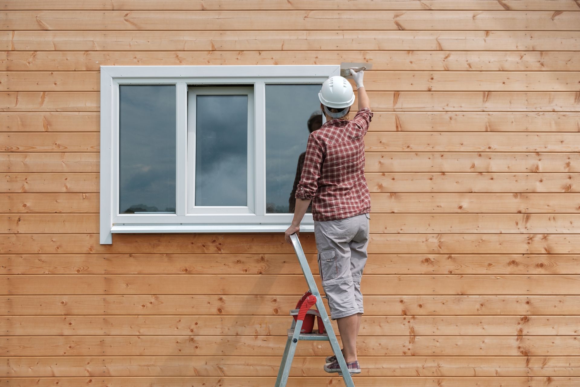 A man is standing on a ladder painting a window on a wooden house.