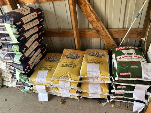 Stacks of animal feed bags inside a wooden barn. Bags are yellow, blue, green, and white.