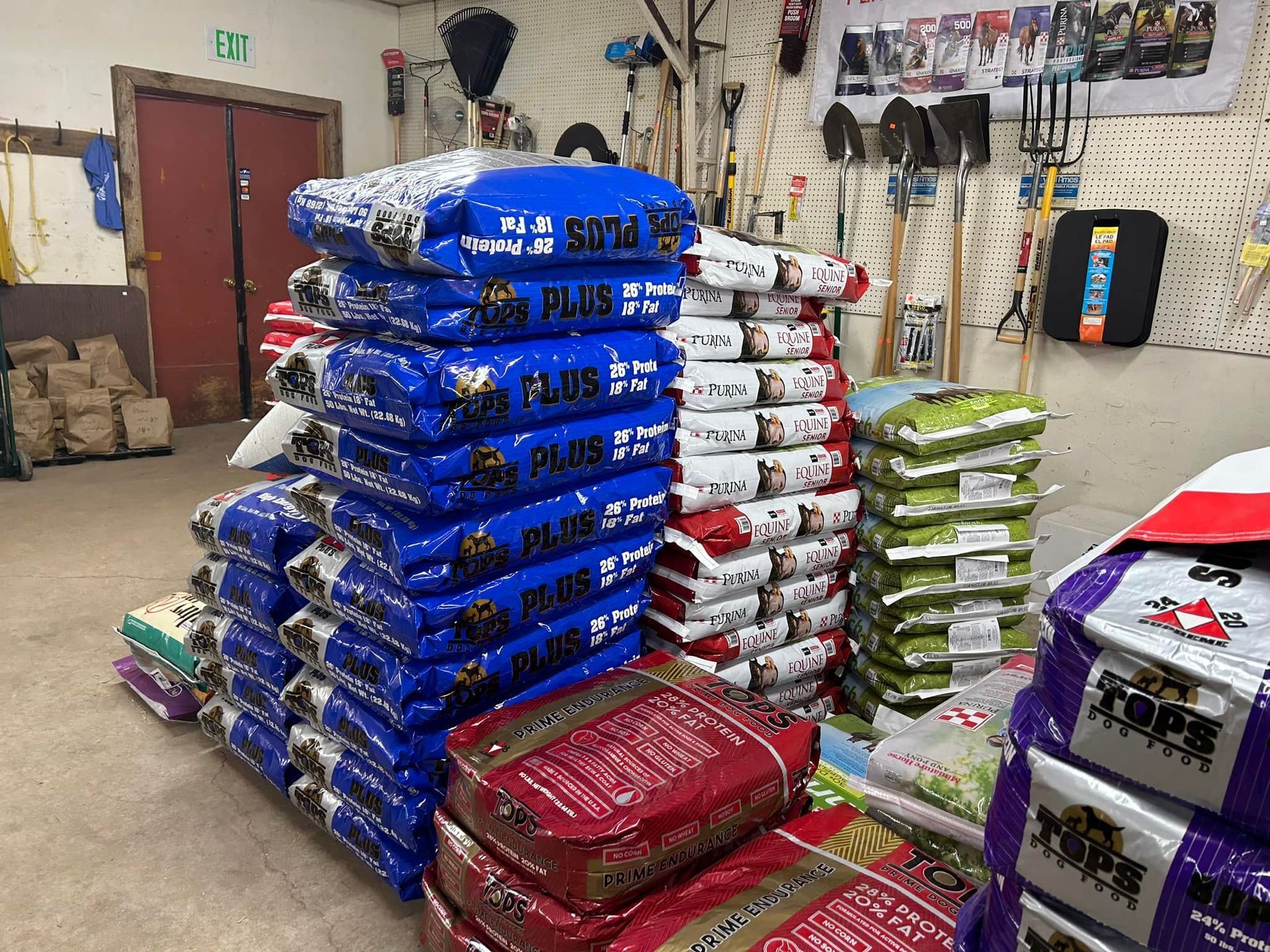 Stacks of feed bags in a store, blue, red, and green bags, tools on wall in background.