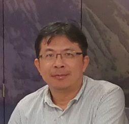 身穿淺藍色條紋襯衫、戴眼鏡的男子麵帶微笑。站在一幅模糊的山巒壁畫前。