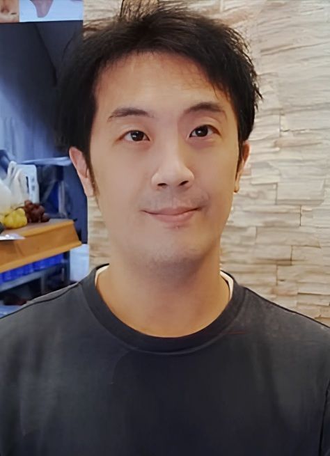 身穿黑色襯衫、黑髮的男子在牆壁和架子前微笑。