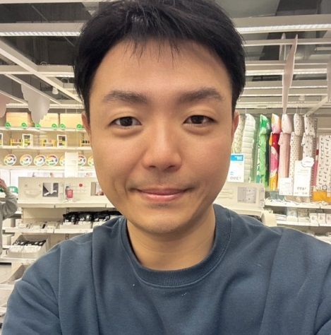 一名男子在室內微笑，可能是在商店裡。他有著深色頭髮，身穿藍色運動衫。他身後是貨架。