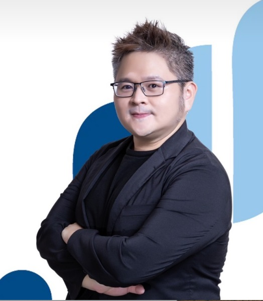 戴眼鏡的男子，雙手交叉，面帶微笑。身穿黑色西裝外套和襯衫。背景為白色和藍色。