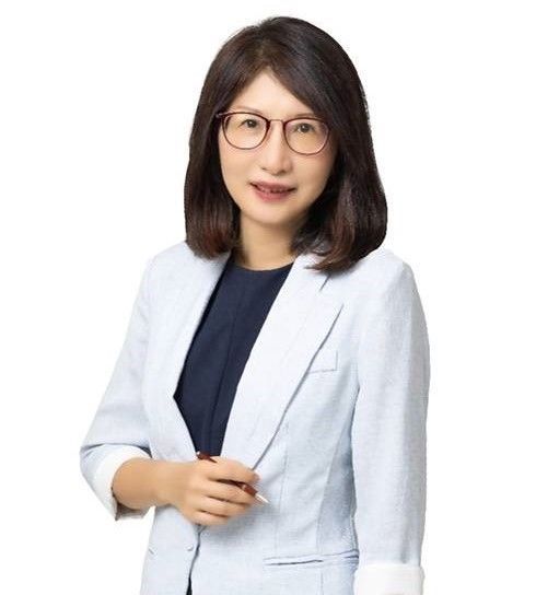 戴著眼鏡、身穿淺藍色西裝外套的女士微笑著，手裡拿著一支筆。