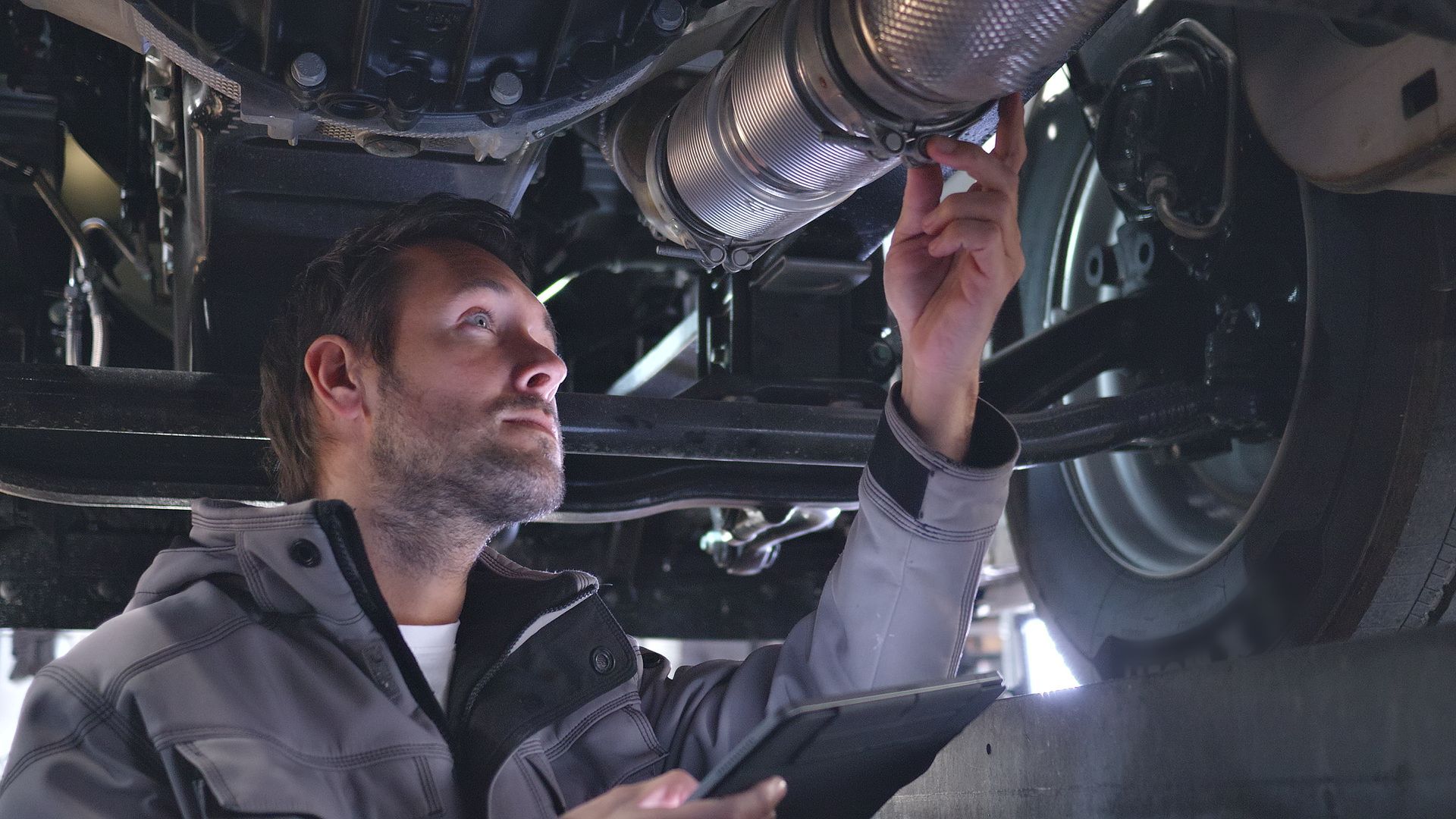 Mechanic inspecting a car's exhaust system. Holding a tablet, looking up at the pipes, under the vehicle.