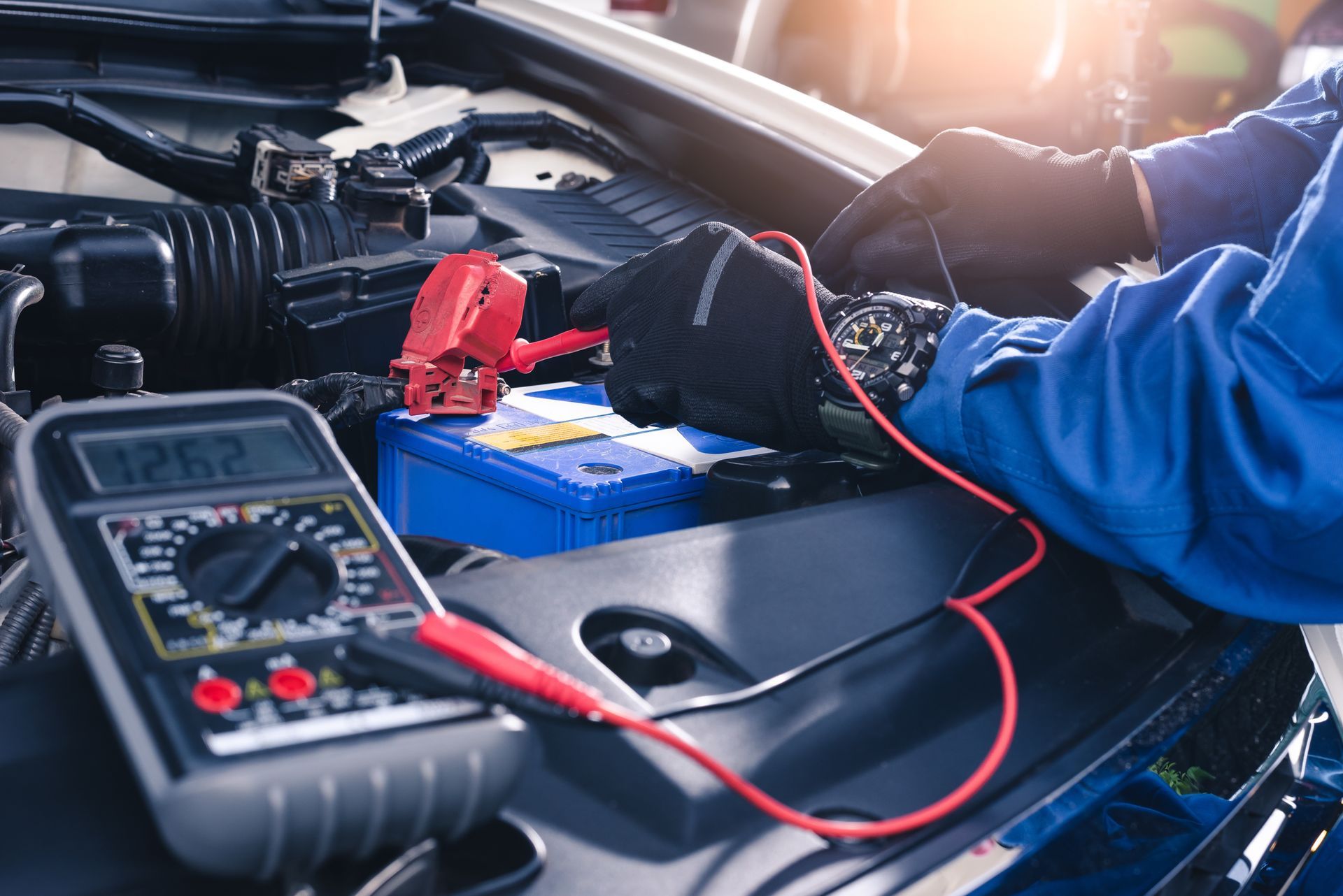 Mechanic using multimeter to test a car battery, inside a vehicle's open engine compartment.