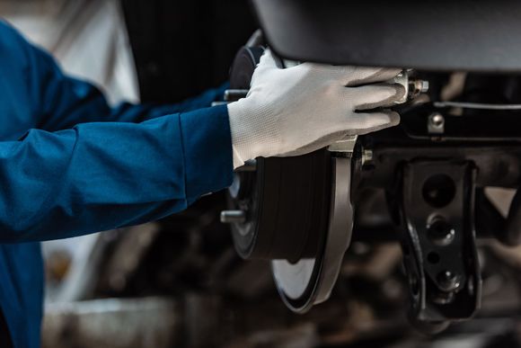 Mechanic wearing gloves working on car brakes; blue jumpsuit, black car parts, shop setting.