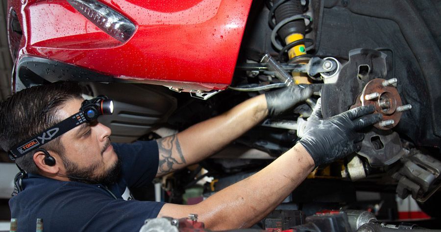 Mechanic repairs a red car's wheel, wearing gloves and a headlamp in a garage.