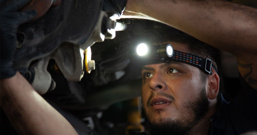 Mechanic working on car, wearing a headlamp. Concentrated expression, hands on vehicle.
