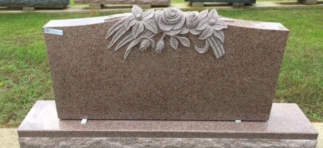 A polished, reddish-brown granite headstone with a carved floral design at the top, set on a matching base in a grassy area.