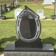 A black granite tombstone shaped like a horseshoe featuring a carved horse head, standing in a grassy cemetery.