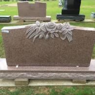 A polished pink granite headstone with a floral carving featuring a central rose, set on a base in a cemetery.