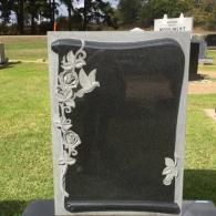 A black upright granite headstone with a carved floral and dove design on the left side, set in a grassy cemetery.