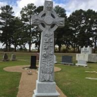 A gray Celtic cross gravestone stands in a cemetery with trees and other headstones in the background.