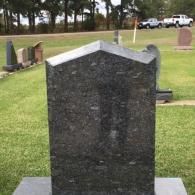 A polished black granite tombstone with a peaked top stands in a green grass cemetery with vehicles visible in the distance.