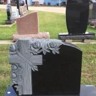A black granite headstone with a carved stone cross and floral detail stands in a monument yard with other markers.