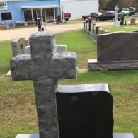 A stone cross monument standing in a grassy cemetery display lot, with a blue building visible in the background.