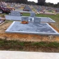 A grey stone headstone on a raised concrete base in a cemetery, with a small wreath of orange flowers in front.