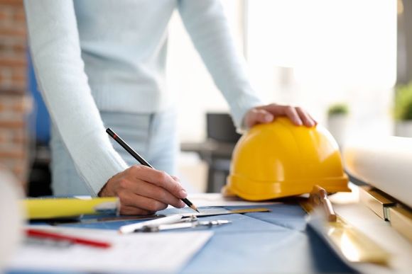 Woman in light blue sweater sketching with pencil, yellow hard hat on desk.