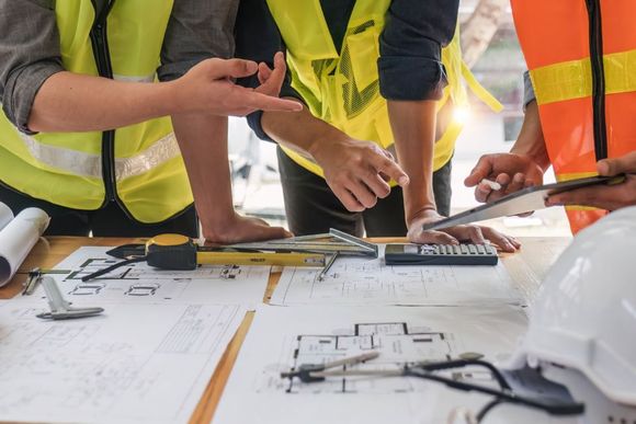 Construction workers review blueprints at a desk with tools, wearing safety vests.