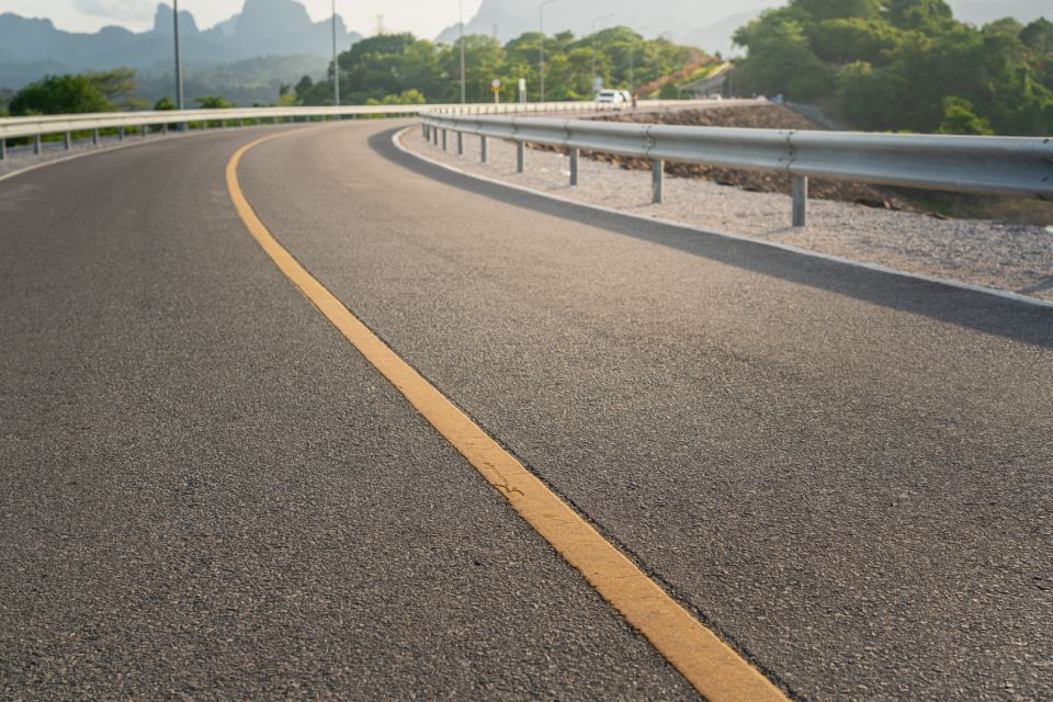 Curved asphalt road with a yellow center line and metal guardrail, mountains in the background.