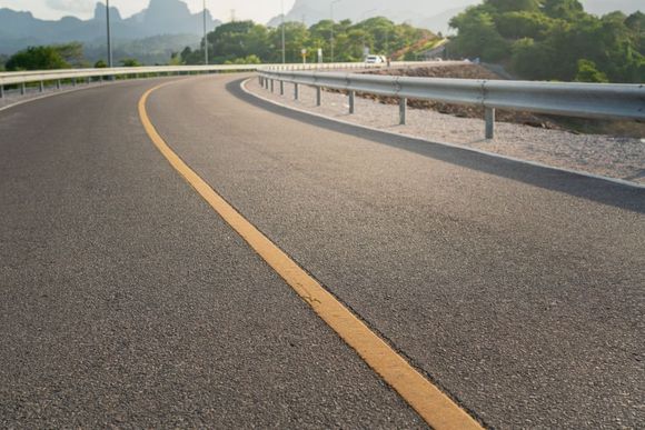 Curved asphalt road with a yellow center line and metal guardrail, mountains in the background.