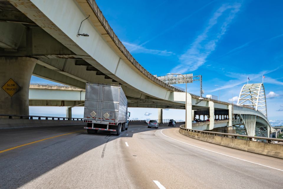 Truck driving on highway under concrete overpass, blue sky in background.