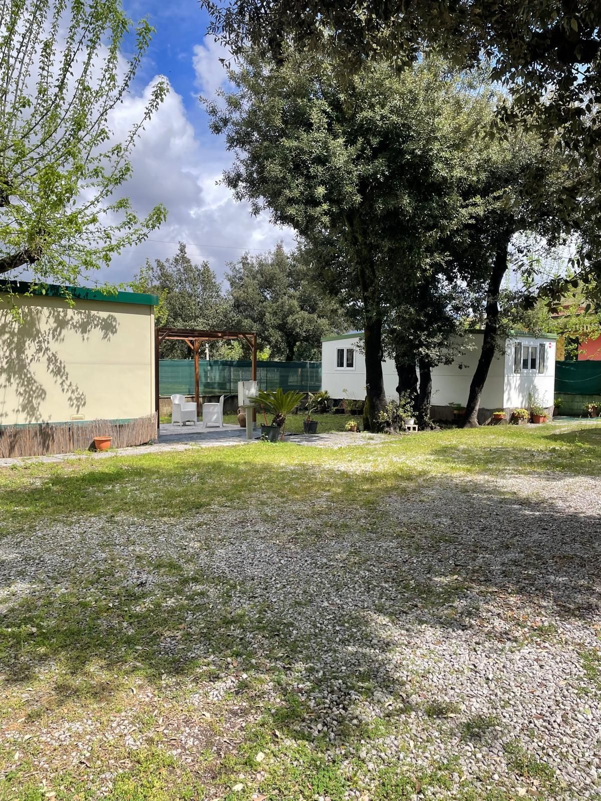 Un cortile ghiaioso che conduce a due piccole case mobili bianche, all'ombra di grandi alberi frondosi, in una giornata di sole.