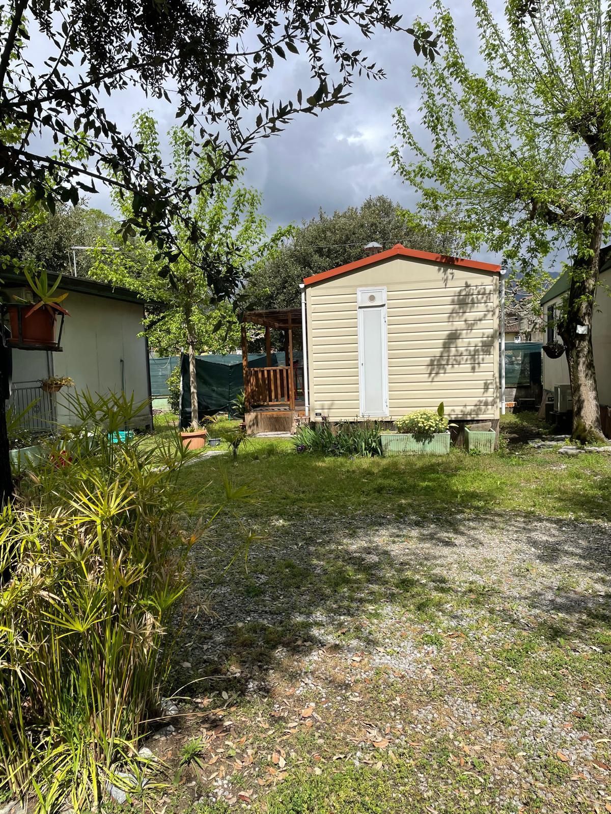 Una casa mobile di colore chiaro con una piccola veranda in legno è situata in un'area giardino con alberi e un cortile ghiaioso.