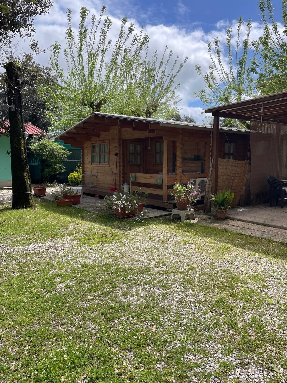 Una casetta di legno con veranda e piante in vaso circostanti, situata in un'area giardino con un cortile di ghiaia e prato.