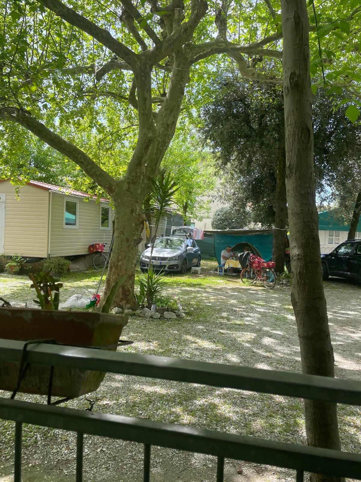 Una vista da una veranda che si affaccia su un campeggio ombreggiato e ricoperto di ghiaia, con una casa mobile, auto parcheggiate e alberi.